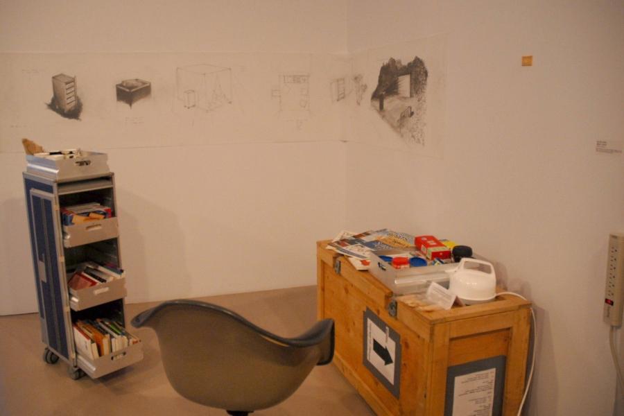 A partitioned corner of the gallery. An airplane snack cart holds biscuit boxes in its top drawer, and books in its middle and bottom drawers. A wooden crate topped with a messy pile of books for learning how to make art, a tray of coffee and tea containers, a tea pot, and a nearly-empty tray of biscuits with an illegible paper sign. A plastic chair sits between them, and conceptual sketches of them are on the partition walls.