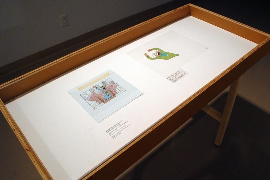 Two stills of cartoon men in coloured sweaters and bowties screaming with their arms raised, displayed in a wood case. The left one depicts its subject sitting in a pink chair at a table, while the right’s subject is drawn on a transparent field.