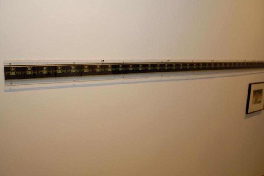 A segment of film rolled out and mounted on the gallery wall, with an indiscernible still mounted to the lower right.