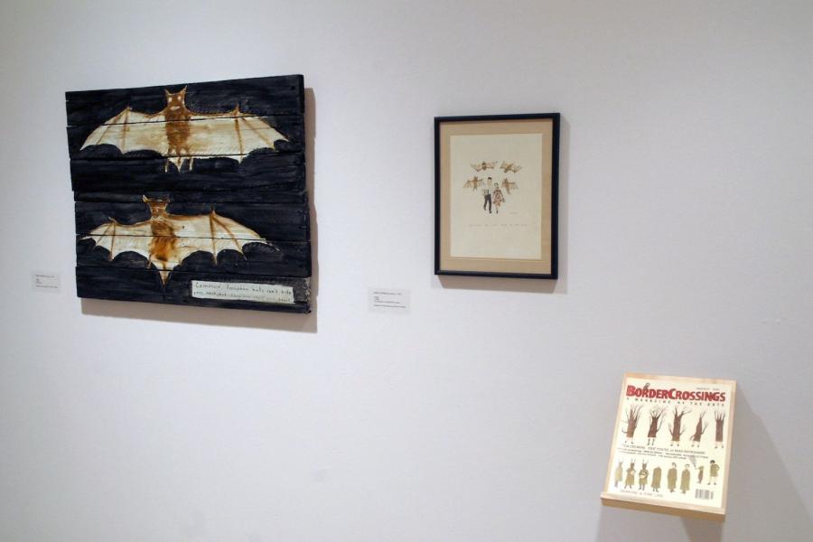 Two framed artworks depicting brown bats and a magazine on a wall. On the left is a painting of two brown bats on black on wooden boards, captioned “Common European bats can’t bite your neck, but they can steal your heart” in the lower right corner. In the centre is an illustration of brown bats attacking a man and a woman, with a small illegible caption. In the lower right is a shelf holding the magazine “drawing a fine line”, with a cover illustration depicting a tree evolving into a human.