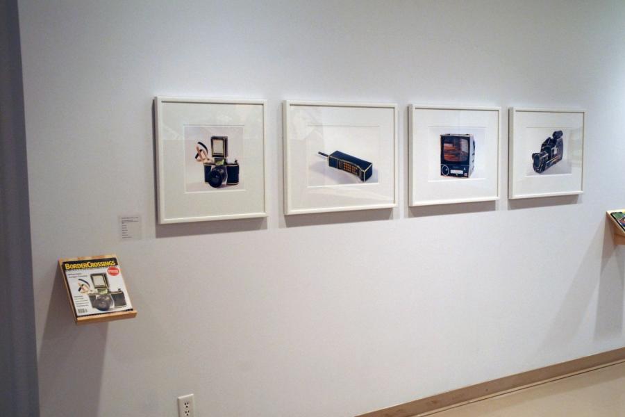 Four framed photos depicting black, yellow-trimmed tech devices and a matching magazine on a wall. From left to right, the framed works depict a camera with a flash and small strap attached, a dynatac phone, a CRT television, and a camcorder. To the lower left, a shelf holds a magazine with the same camera on its cover.