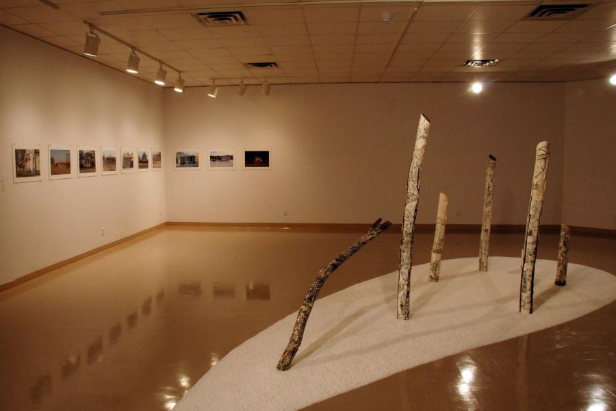 The gallery viewed from above. A large sculpture depicting seven birch tree trunks chopped to various heights stands on a white surface in the centre of the room, resembling snow. One trunk is only partially visible behind another.  And in the back corner, ten street photographs are hung in a line on the wall.