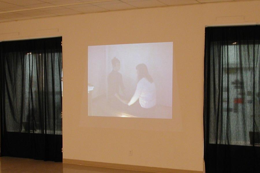A video projected on the gallery wall with translucent black curtains covering the windows on each side, and a bench in front. The captured still features two people sat facing each other and holding hands.