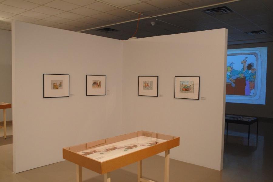 The gallery. There are four animation stills hung on a detached wall in the centre, four stills of a boat lined up in a wooden display case in front, and a cartoon animation projected onto the very back wall behind them.