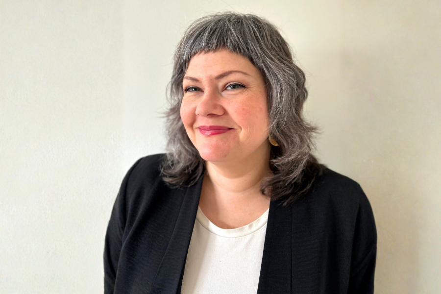 Portrait of Lisa Wood with short, layered grey hair smiling softly at the camera, wearing a black blazer over a light-coloured top, photographed against a plain white background.