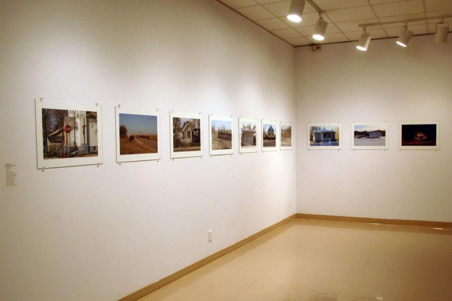 Ten street photographs hung in a line along the corner of two walls. a street sign reading “logan ave” in front of a white church, a road crossing over a creek surrounded by dead plants, a white house, a tree-lined dirt path, a blue house, a yellow house, an orange corner store, another white house, a fallen letter board, and a Christmas light display at night.