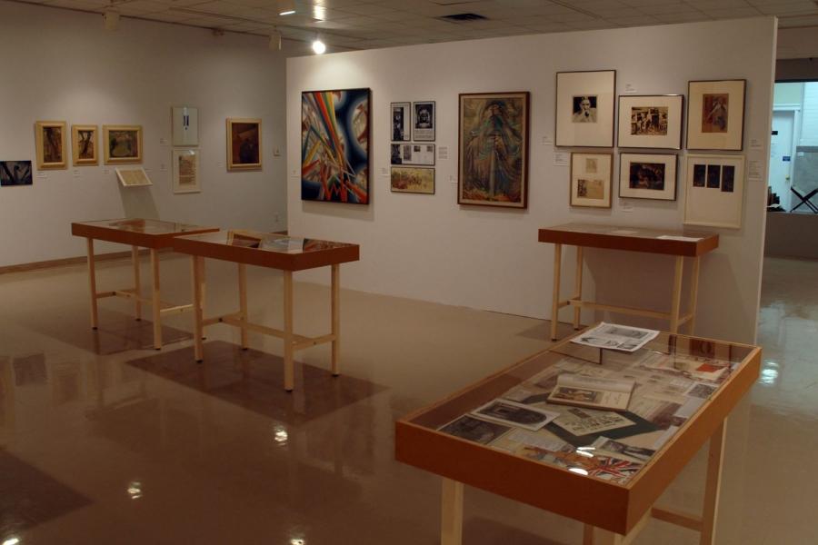 A view of the gallery including paintings on the walls, archival documents in display cases, and books in the far windowsill. On the central partition wall is a painting of multicolored tubes in a grid of blue and red circles, a painting of a tree in circular brushstrokes, a black and white portrait of a man, a black and brown sketch of grain elevators, and other indiscernible works.
