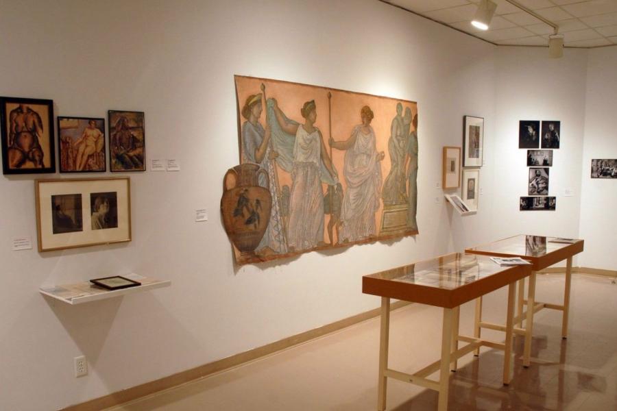 A gallery wall and two display cases of archival materials. From left to right, the wall includes: three artworks of a nude woman paired with two photographs of a clothed woman and a shelf of indiscernible pieces. Then a large painting imitating a Greco-Roman fresco featuring figures in togas, a red-figure vase, and a statue. Then three indiscernible works hung in frames, with another on a shelf. Then five indiscernible black and white photographs.