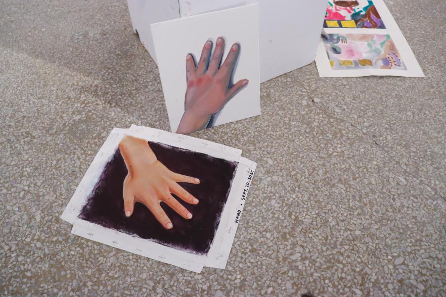 Painted studies of hands are arranged on a speckled studio floor, including a realistic hand on dark ground and a smaller panel leaning upright.