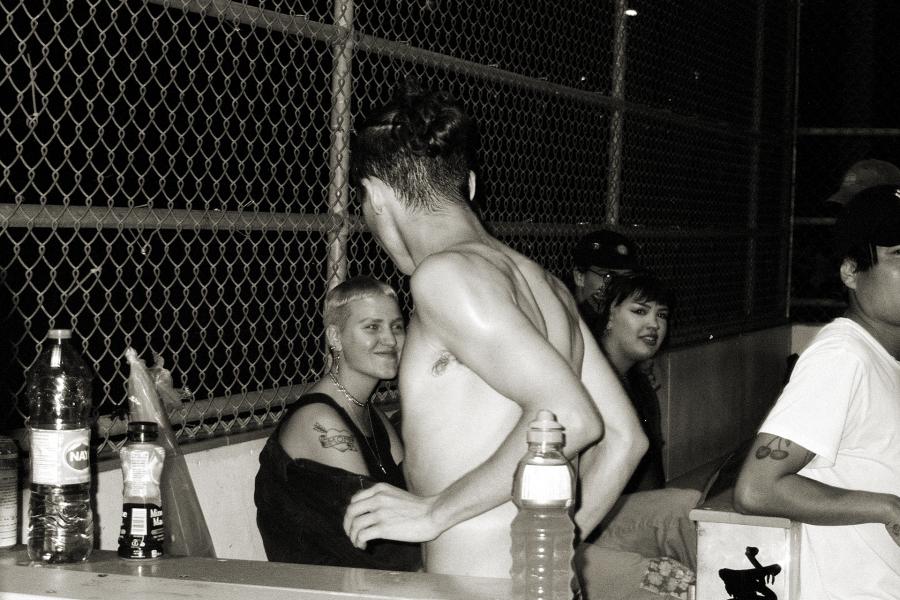 Black-and-white photograph of several people gathered beside a chain-link fence at night, with one shirtless figure standing in the foreground while others sit and talk nearby.