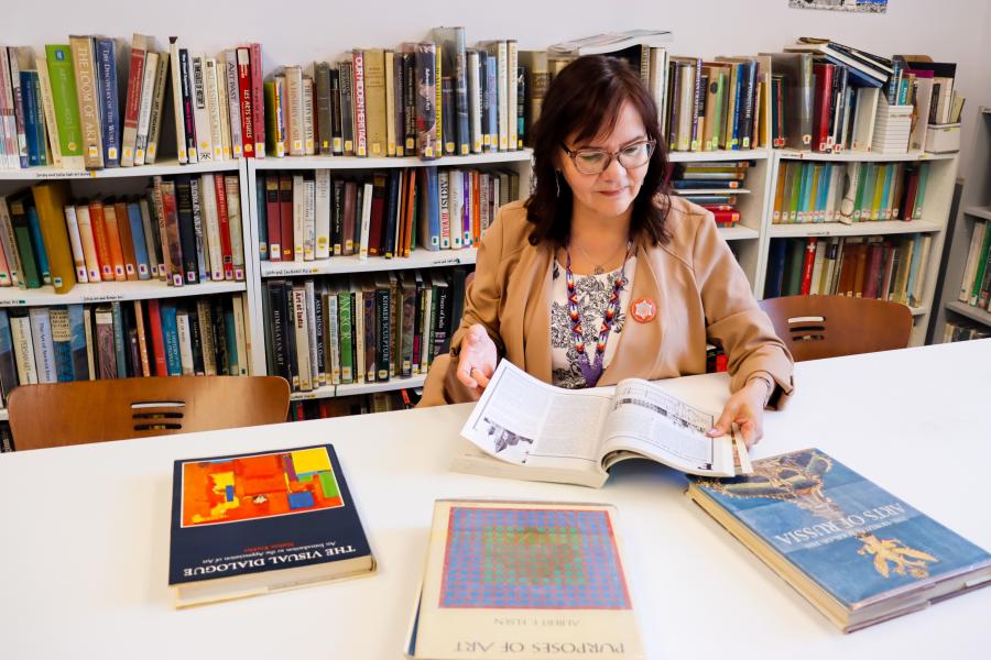 sits at a table in front of bookshelves, reading an art book in a library setting.