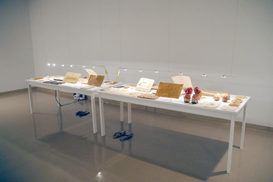 Installation view of the BorderCrossings Study Centre exhibition featuring two display tables with artist books, sculptures, and mixed-media works illuminated by suspended task lights.