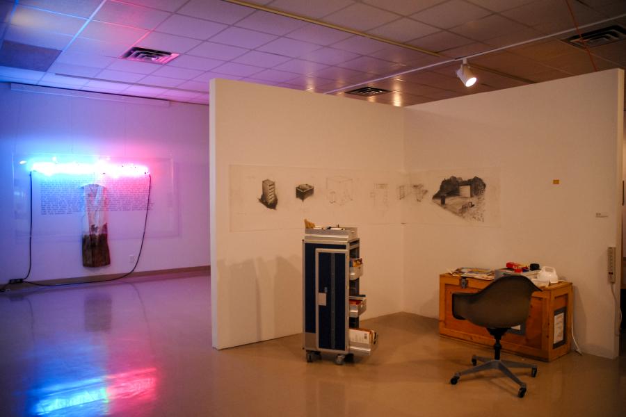 Installation view of a contemporary art exhibition featuring a workspace with a desk, chair, and rolling cabinet, charcoal drawings on partition walls, and a neon light installation illuminating the gallery in the background.