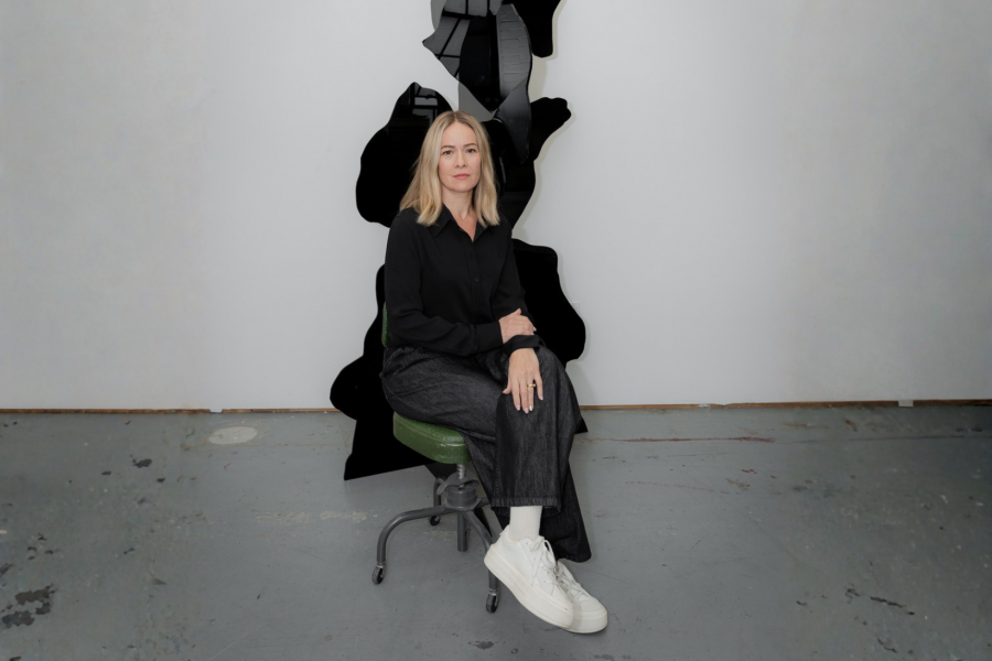 Portrait of artist Dominique Rey seated on a green rolling stool in a studio or gallery space. She wears a black shirt, dark jeans, and white sneakers.