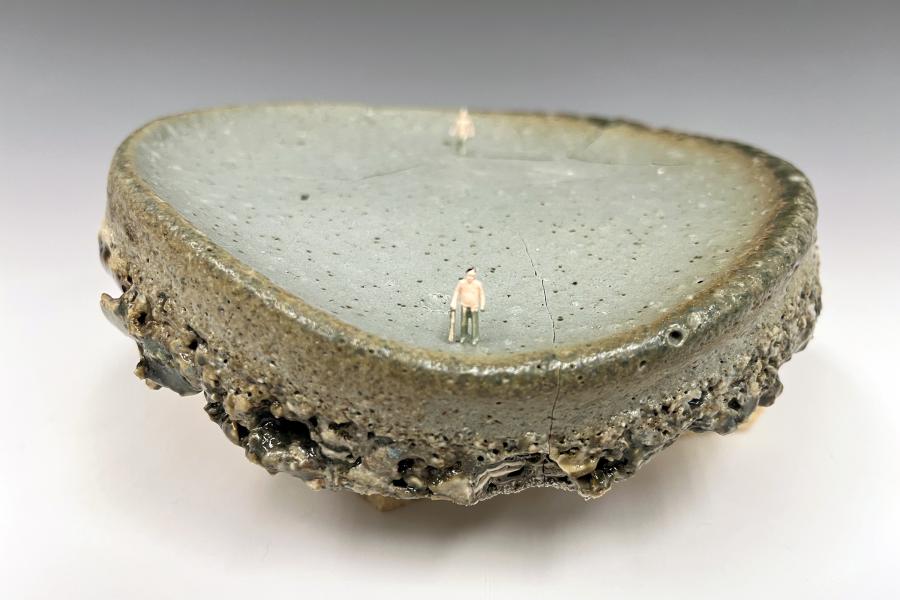Ceramic disc by Lin Xu with a pale grey-green, pitted surface ringed by a thick, rough, glazed edge that looks like layered rock. Two tiny human figures stand on the surface, one in the foreground holding a cane and one in the distance.