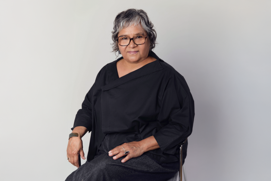 Portrait of Lori Blondeau seated on a chair, wearing tortoiseshell glasses, a loose black top, a metal cuff bracelet and rings, against a light gray background.”