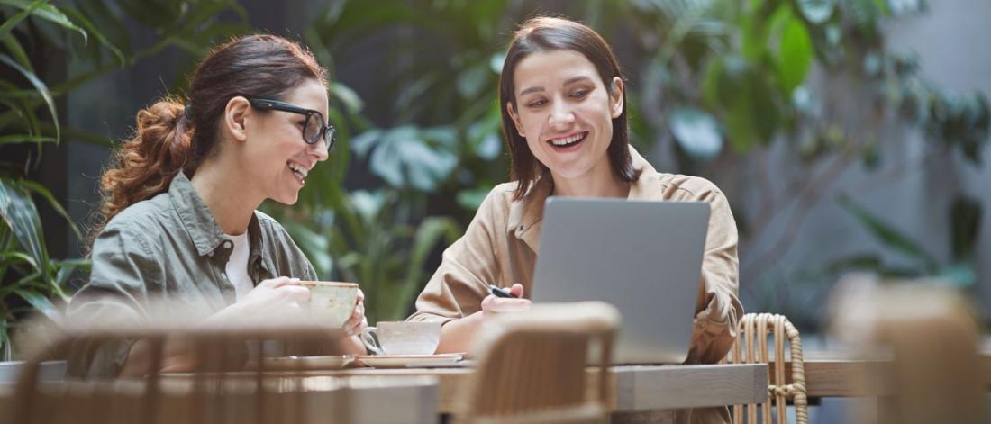 Two students sitting at long table with coffees looking at laptop.