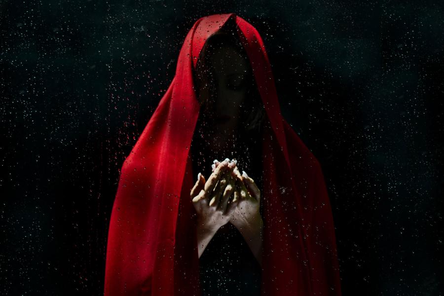 Red cloaked figure emerging from the dark with hands reaching outwards.
