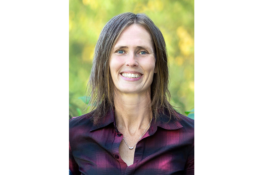 Headshot of smiling woman wearing a purple plaid shirt with a background of trees.