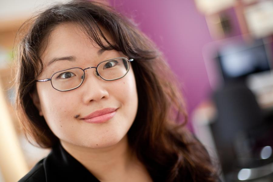 Headshot of woman with long dark hair and glasses in front of a purple wall backdrop.