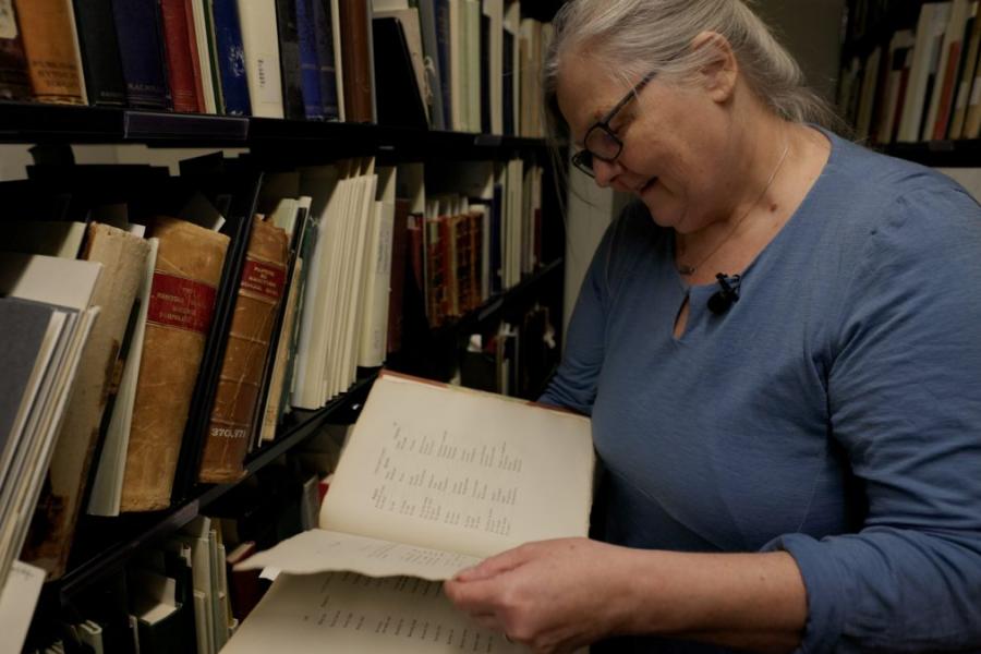 Anne Lindsay looking down at an open book standing in front of a library bookshelf full of books and reports.