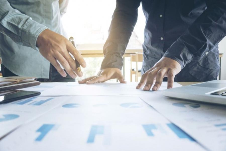 A close up of the hands of two males looking over a number of printed graphs.