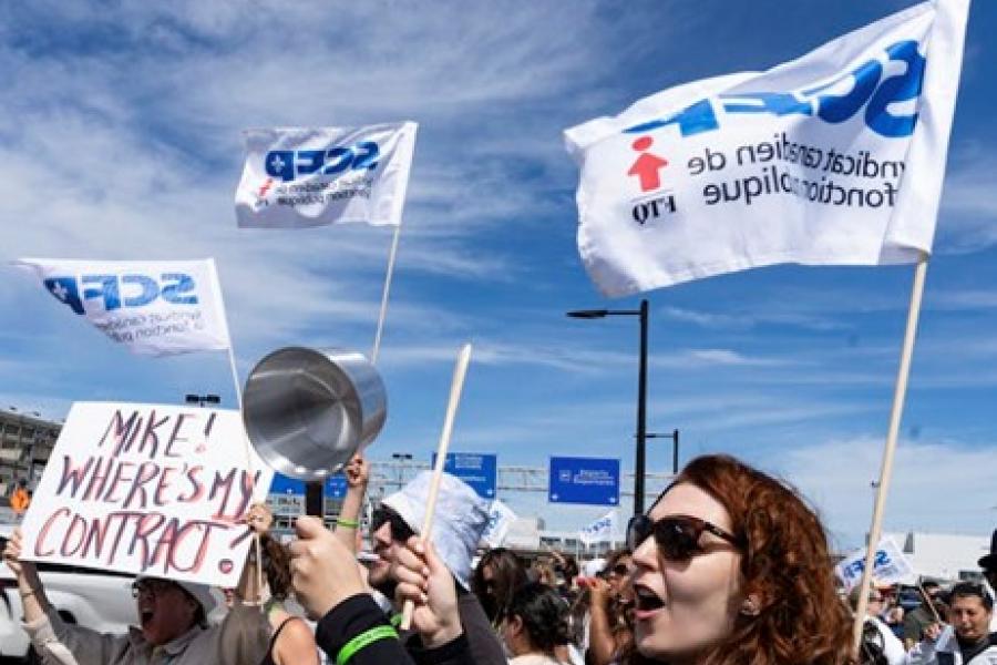 A crowd of people walking in an outdoor protest, holding up signs and banging pans. 