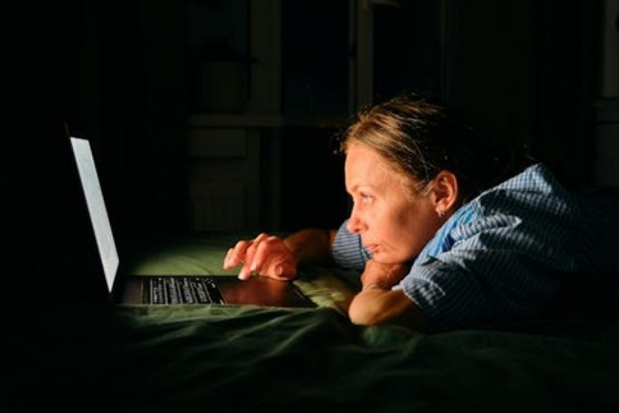 A woman lying stomach down on a bed, in the dark, staring into a laptop screen.