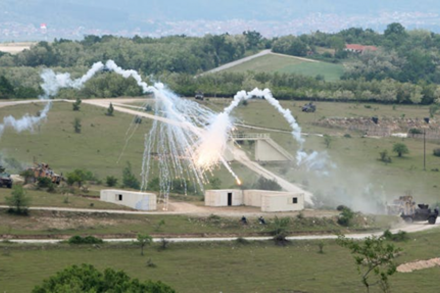 A field with some outbuildings with military bombs exploding during an exercise.