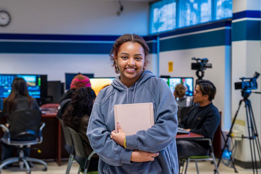 A happy student inside the media lab holding a laptop and standing in front of cameras and podcast table 