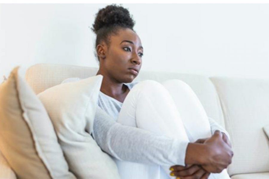 Woman sitting on a sofa with her knees pulled close and arms wrapped around her knees. She has a sad look on her face.