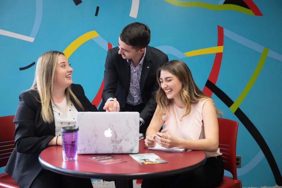 Three smiling people sit at a red table in a bright blue room.