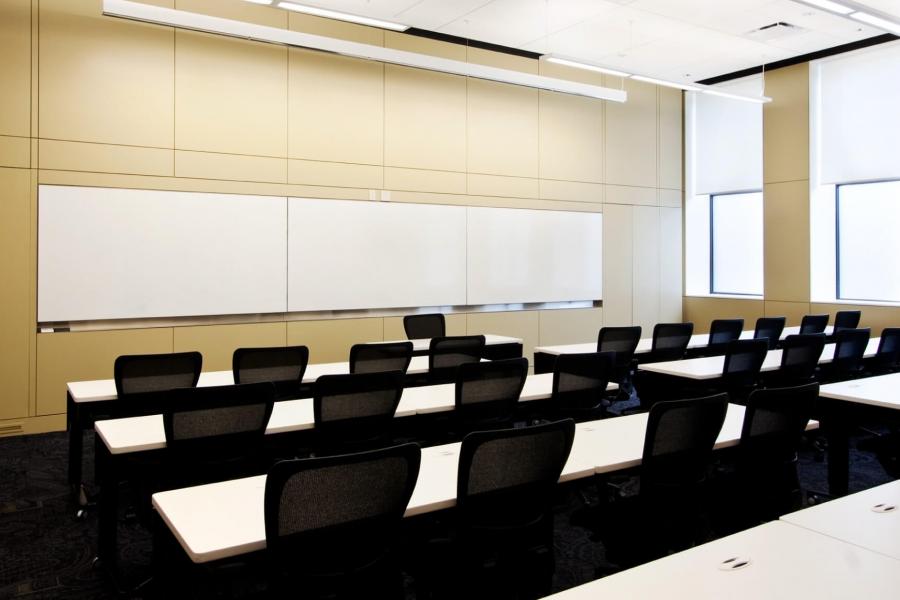 A large classroom with several narrow rows of tables and chairs and  a large whiteboard at the front of the room. 