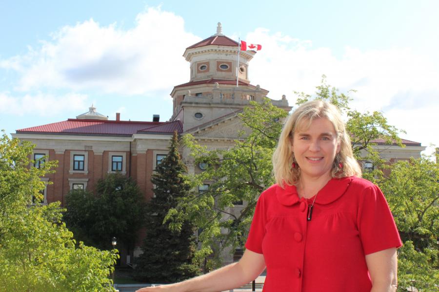 photo of Kelley Main wearing a red shirt. the university of Manitoba administrative building is in the background