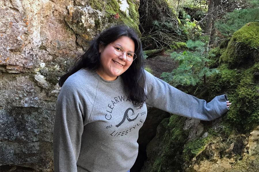 Young woman, Jennifer Moose, wearing a grey crew neck sweater standing with her hand a boulder with rocks and trees around her.