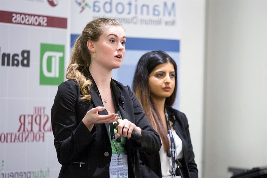 Two women standing up talking to an unseen audience, both wearing blazers. One of the woman is blonde and is deep into her speech.