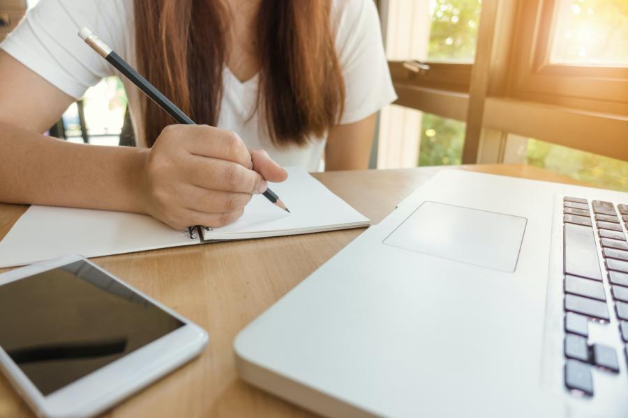 Woman with long hair wearing a white shirt writing in a notebook behind a laptop.