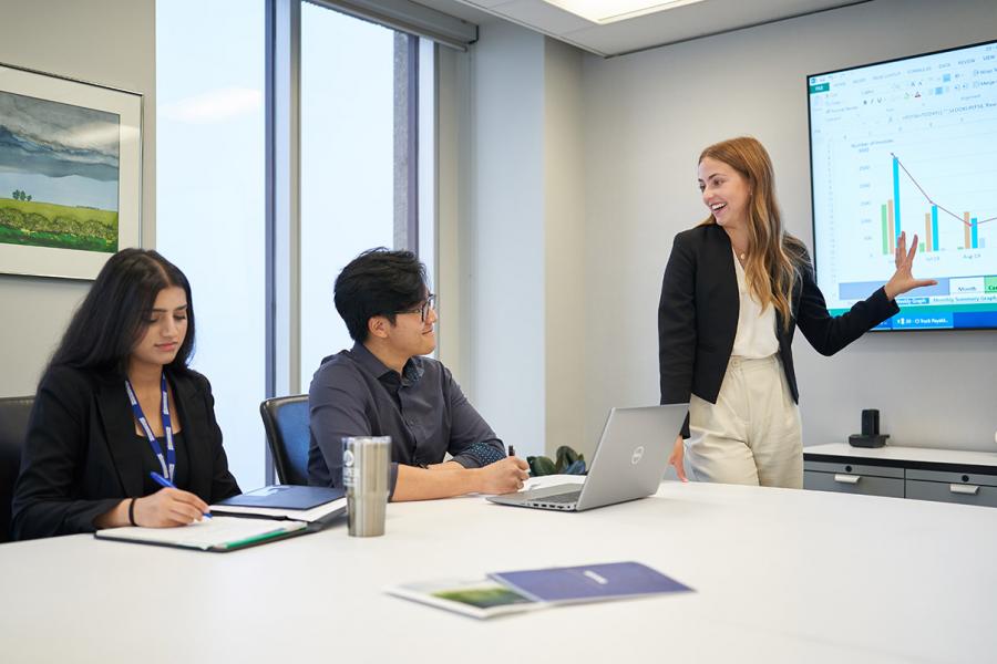 Three Asper School of Business students in a boardroom, one is presenting and looking at the other two who are sitting down. 