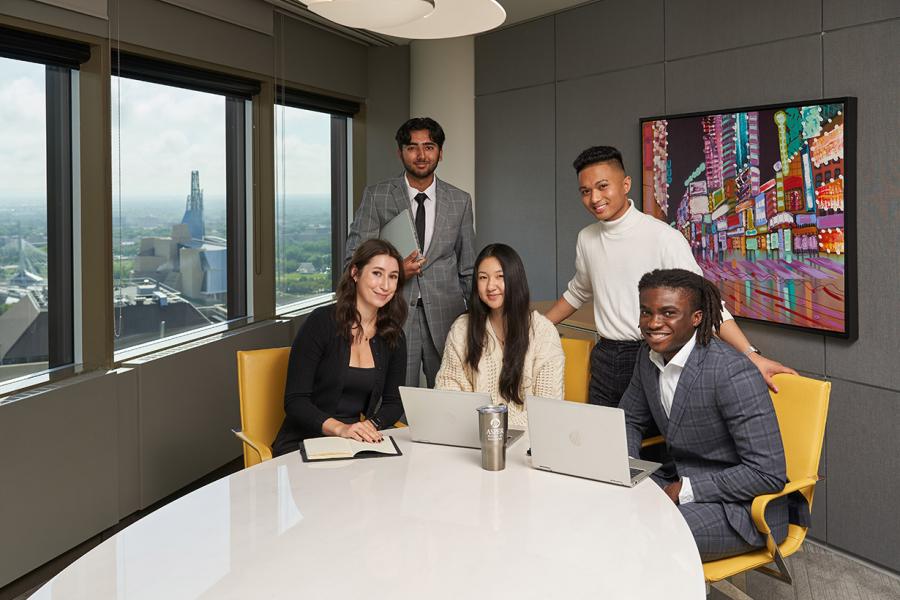 Five students wearing business clothes in a grey boardroom. The Human Rights museum can be seen behind them through the window.