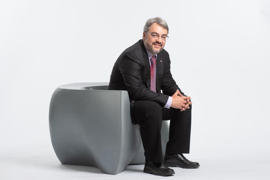 Bruno Silvestre wearing a black suit sitting on a modern grey chair smiling. 