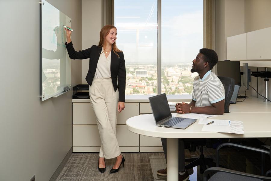 Female student writing on a whiteboard while a male student sits a desk.