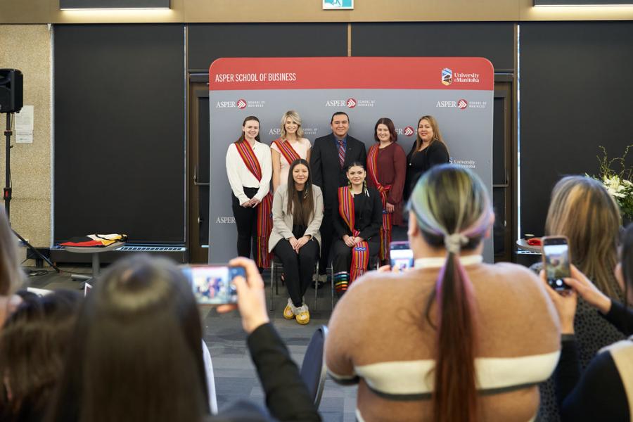 Group of Indigenous Asper students standing behind a backdrop with people taking pictures of them.