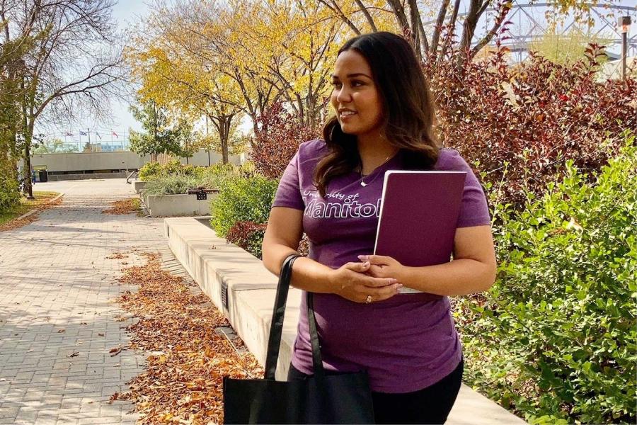 A student wearing a maroon University of Manitoba t-shirt, holding a tote bag and notebook.