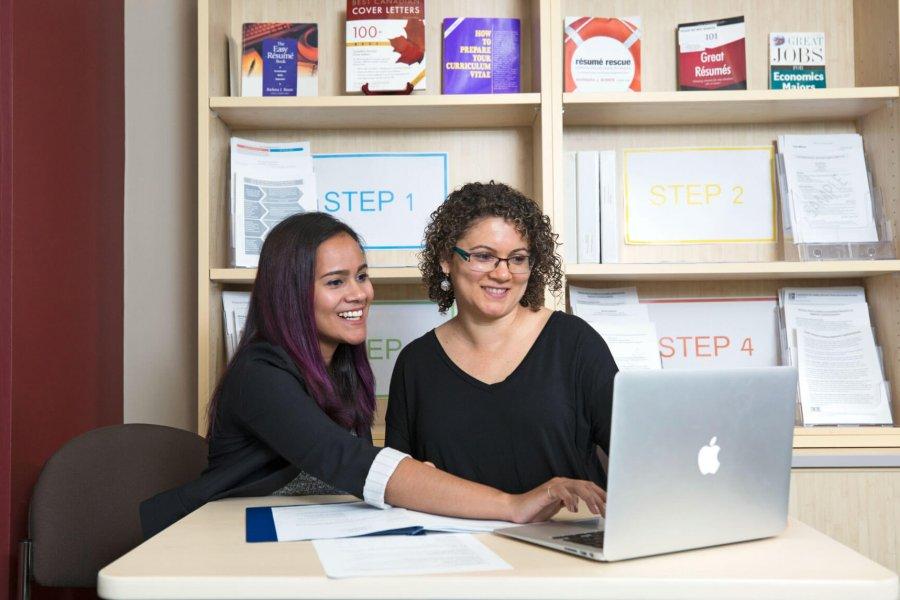 A Career Services employee offers assistance to a student while they sit at a desk together in front of a laptop. 