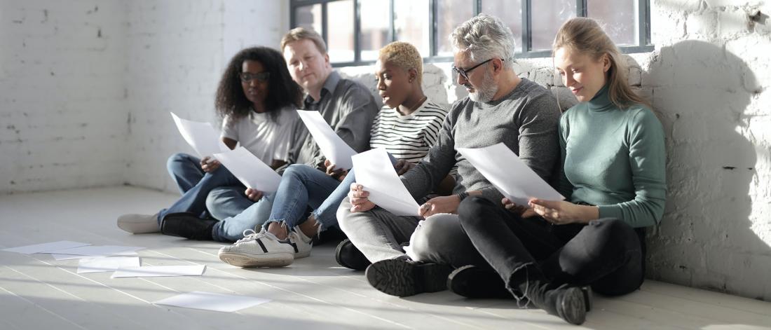 An intergenerational group of five people of different ages sitting on a wooden floor by a window reading papers in a group.