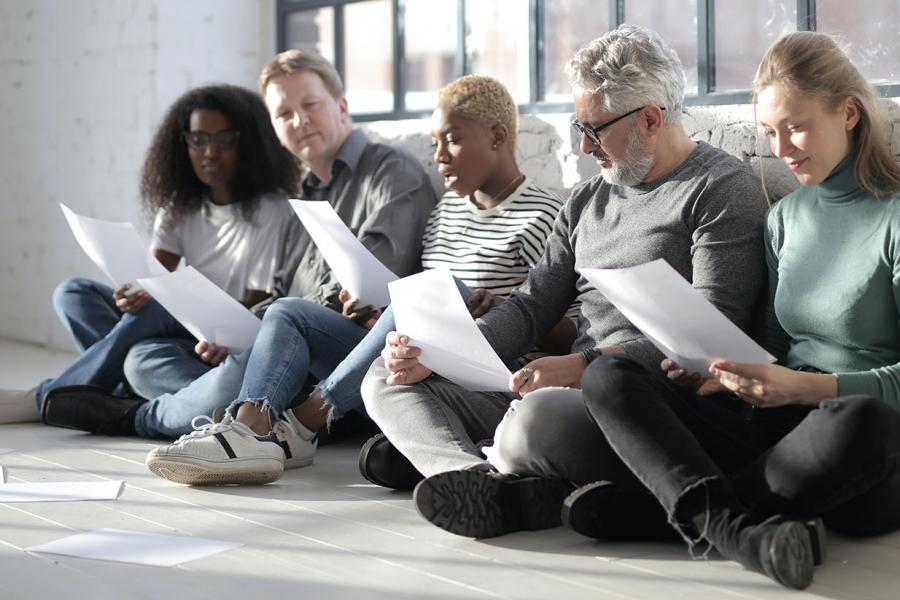 An intergenerational group of five people of different ages sitting on a wooden floor by a window reading papers in a group.