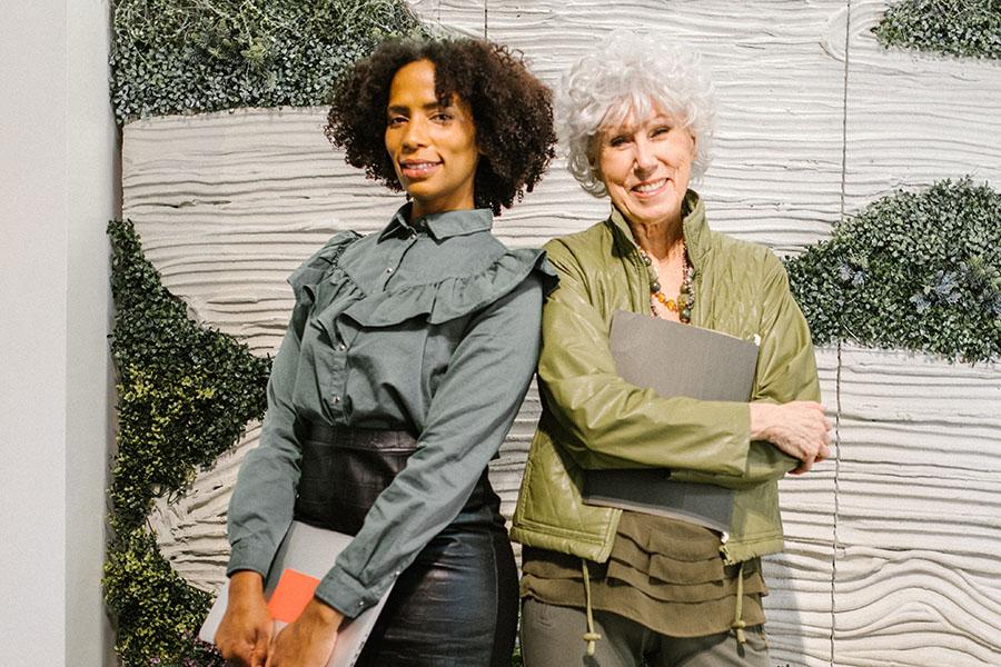 Two diverse women, who are different ages are smiling and standing back to back.