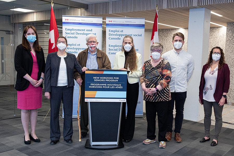 Seven University of Manitoba and senior organization community members stand between two banners and a podium at the New Horizons funding announcement.