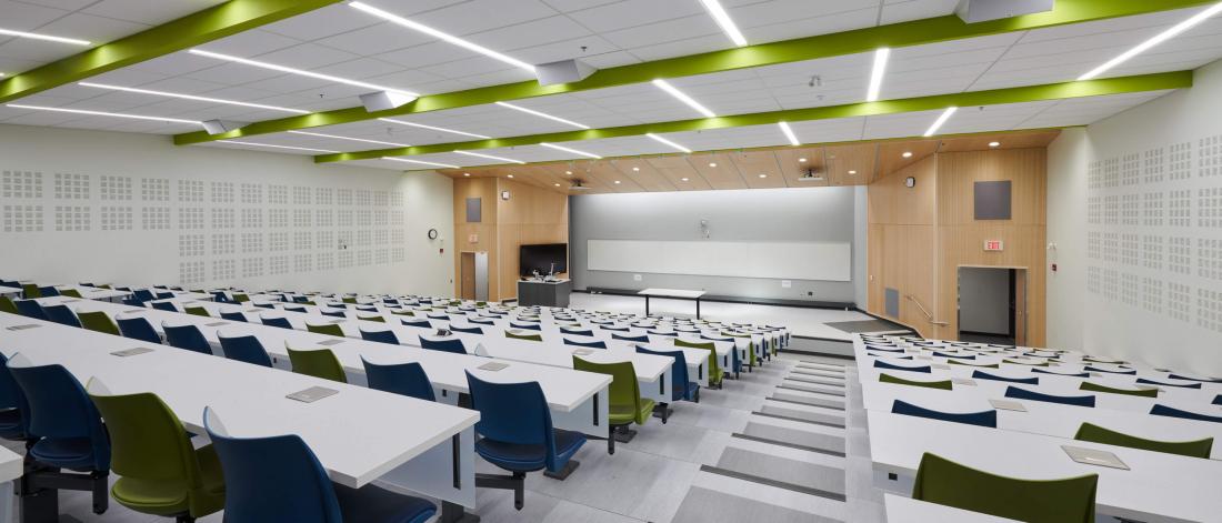 The brightly lit modern classroom space of room 290 from the back row looking towards the front of the room with rows of bright white tables and green and blue chairs.