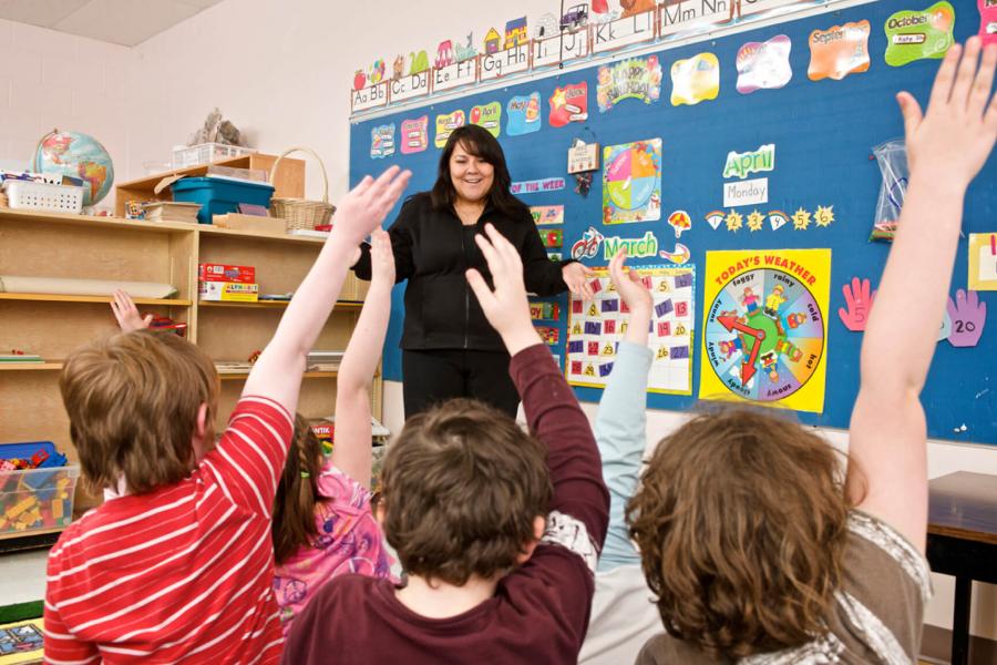 A smiling teacher stands in front of an elementary grade classroom full of excited students with raised hands. 